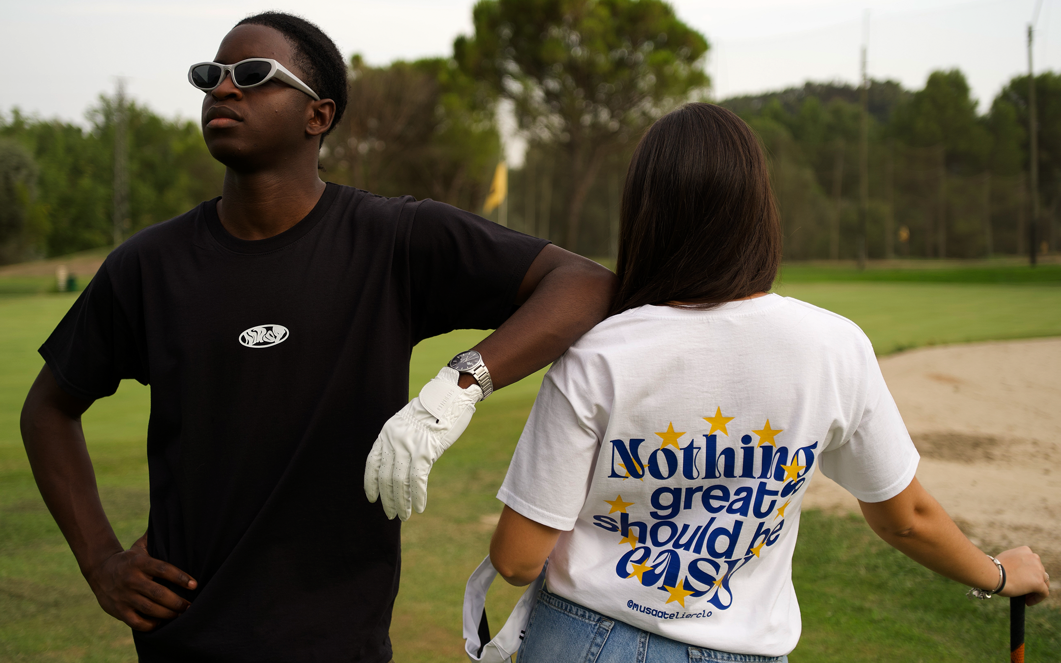 A man in a black t-shirt and white sunglasses stands next to a woman on a golf course. The woman wears a white t-shirt with blue and yellow text that reads 'Nothing great should be easy' and 'Bourgeois Lifestyle' at the bottom.