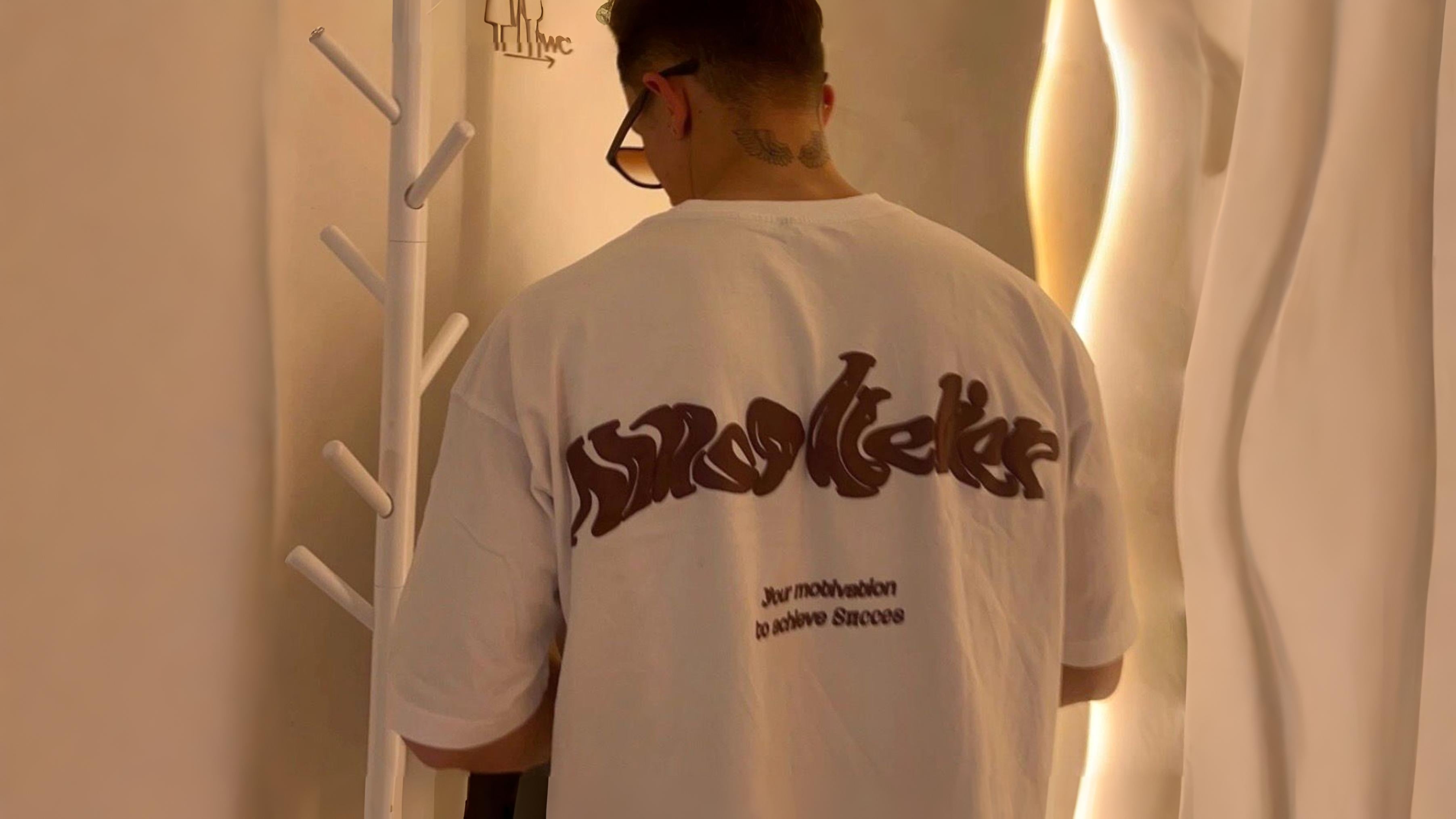 Person wearing a white t-shirt with text on the back, standing in a minimalistic room.